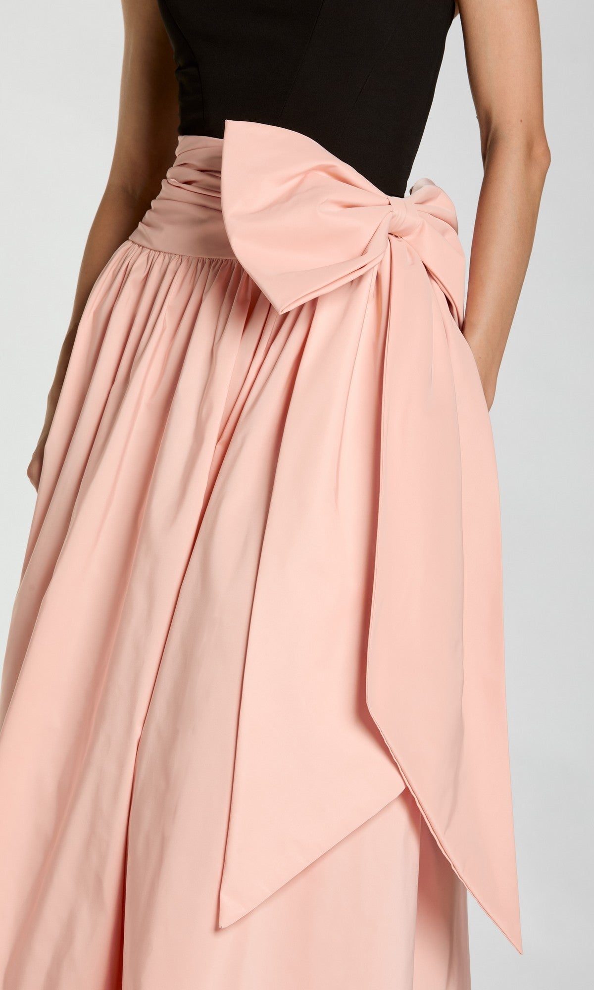 A person wearing the Mac Duggal 2370 long formal dress, featuring a black top and a light pink oversized bow at the waist, shown from shoulders to knees.